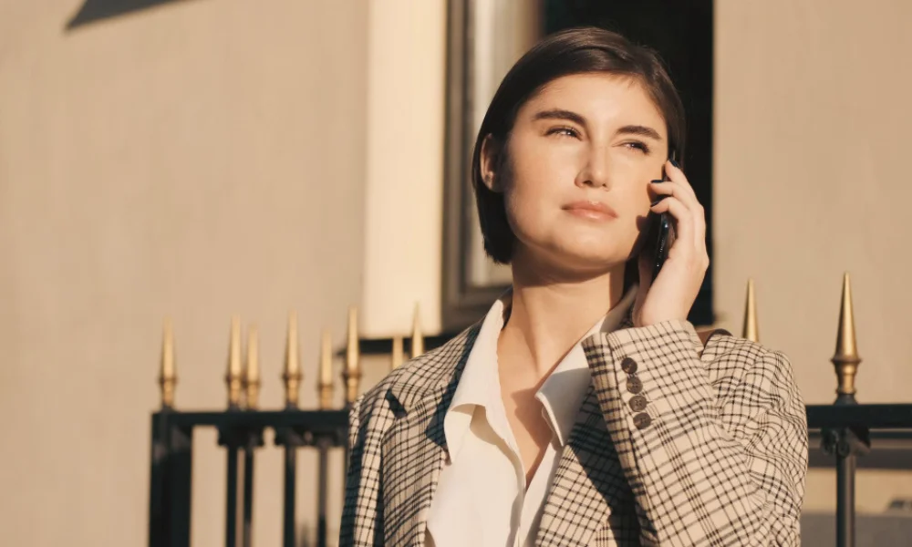 A woman with short hair in a plaid blazer is talking on the phone outside in sunlight
