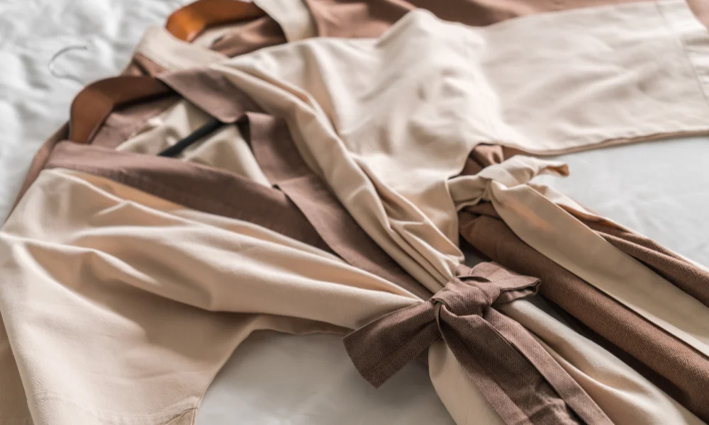 A beige and brown robe with a tied belt is neatly placed on a white bed