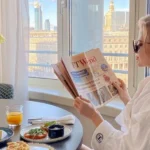 Woman in robe reading newspaper by window with breakfast