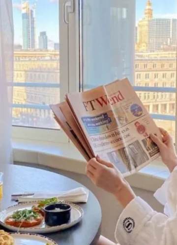 Woman in robe reading newspaper by window with breakfast