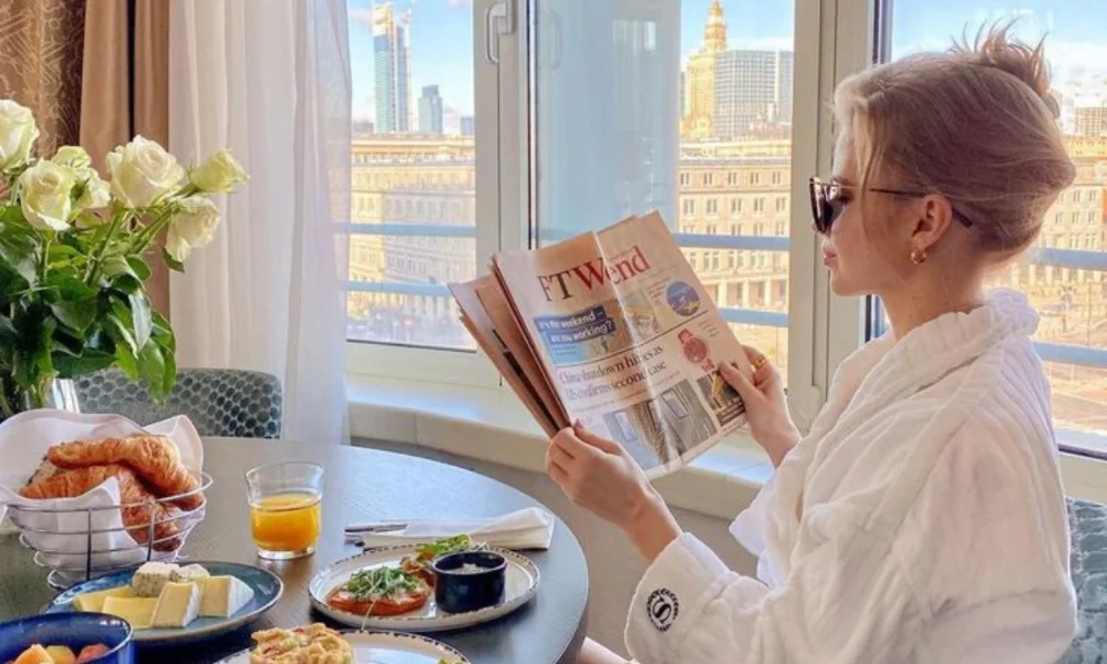 Woman in robe reading newspaper by window with breakfast