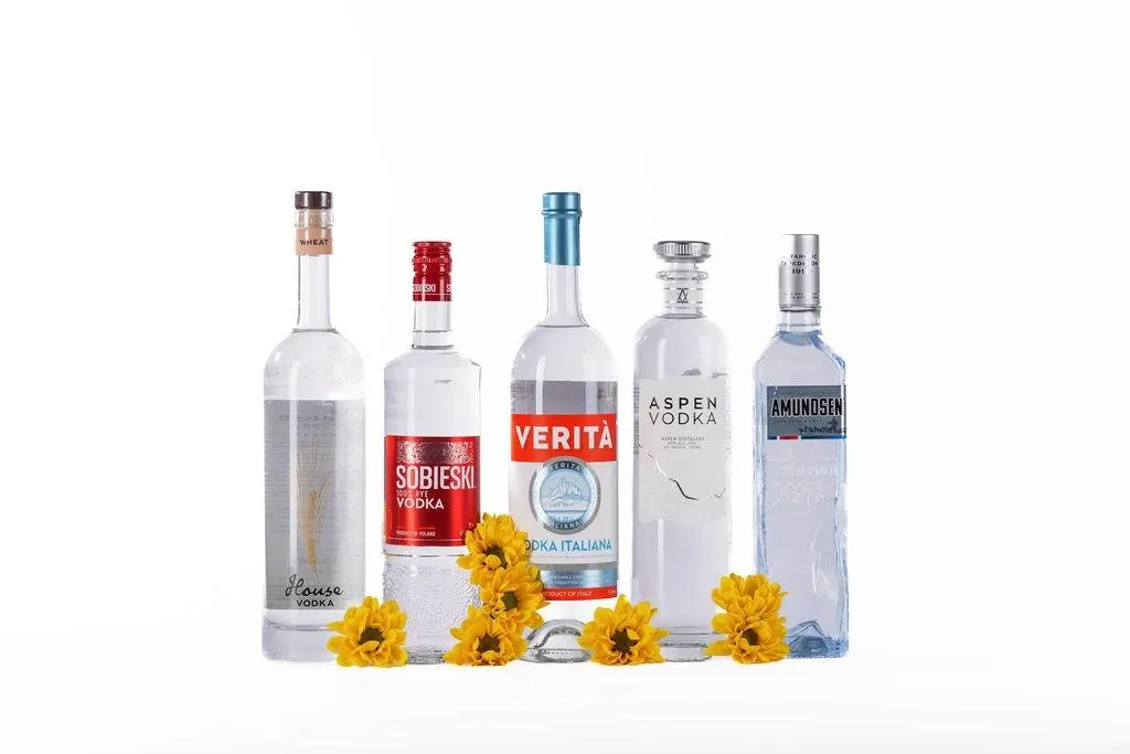 Five different bottles of vodka displayed with yellow flowers on a white surface.