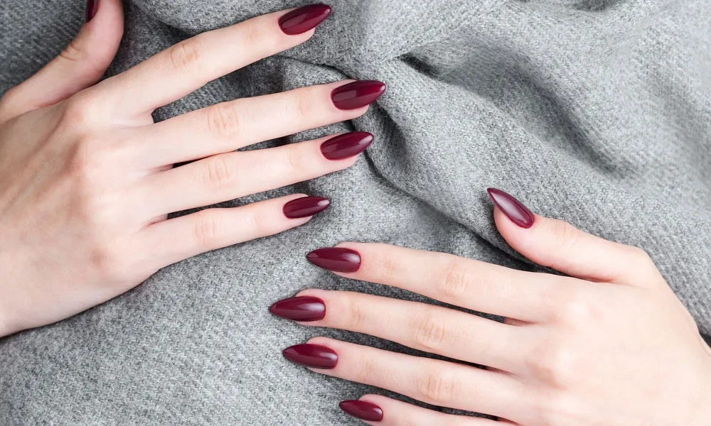 Two hands with glossy burgundy almond-shaped nails rest on soft gray knit fabric, highlighting a polished manicure in an elegant close-up.