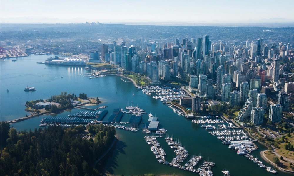 Aerial view of Vancouver’s dense glass-and-steel skyline beside a blue harbor, with marinas and forested waterfront in bright, calm daylight.
