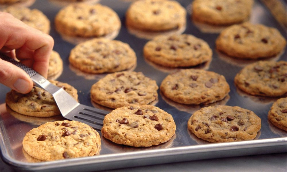 A hand slides a metal spatula under a golden chocolate chip cookie on a tray crowded with freshly baked cookies, creating a warm, inviting scene.