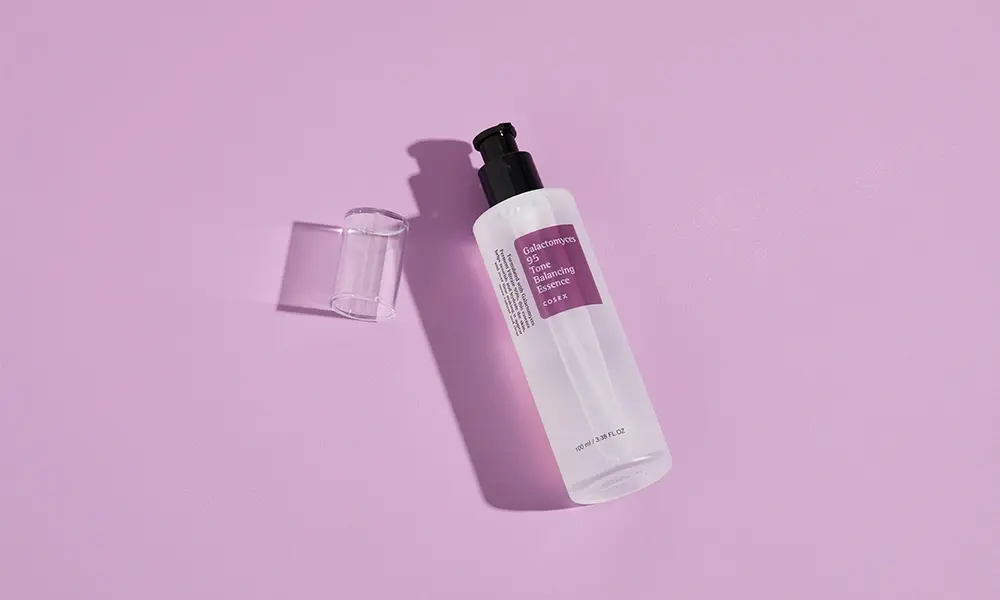 A clear skincare bottle with a black pump and purple label lies beside its transparent cap on a pink surface, casting crisp shadows for a minimalist, serene look.