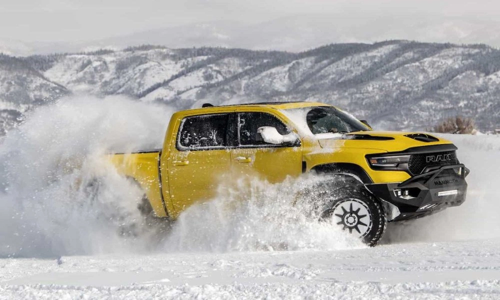 A yellow Ram pickup powers through deep snow, spraying powder around its tires, with snowy mountain ridges behind it; a rugged, adventurous scene.