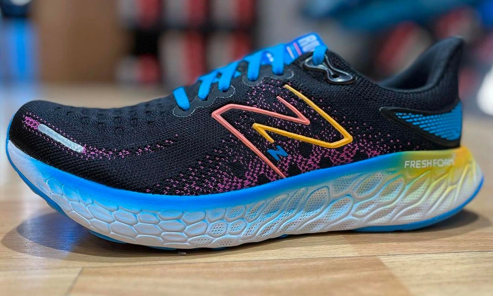 A black running shoe with bright blue laces, pink accents, and a yellow logo sits on a wooden surface; its thick blue-white sole suggests cushioned performance.