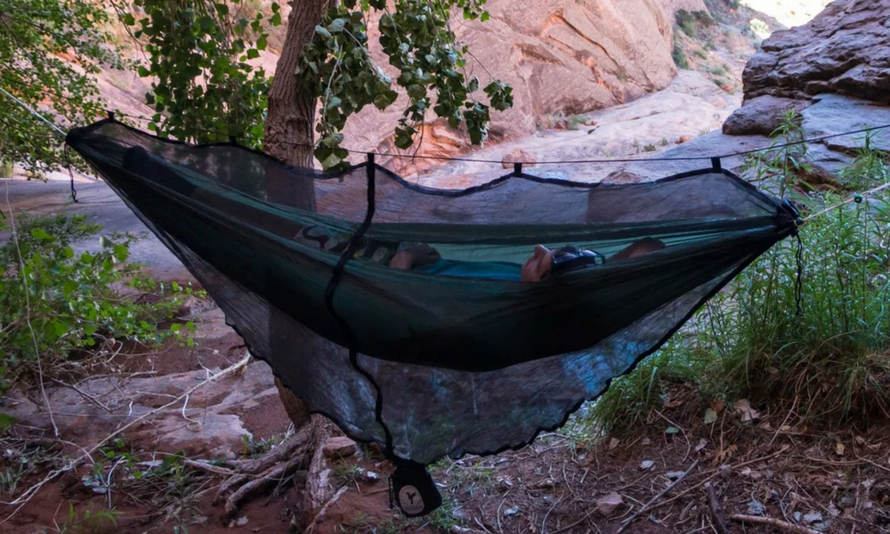 A person lying down inside a green camping hammock enclosed by a black mesh bug net
