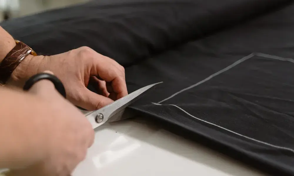 Hands cut dark fabric with metal scissors along a chalk guideline on a worktable, capturing a focused, careful moment in garment making.