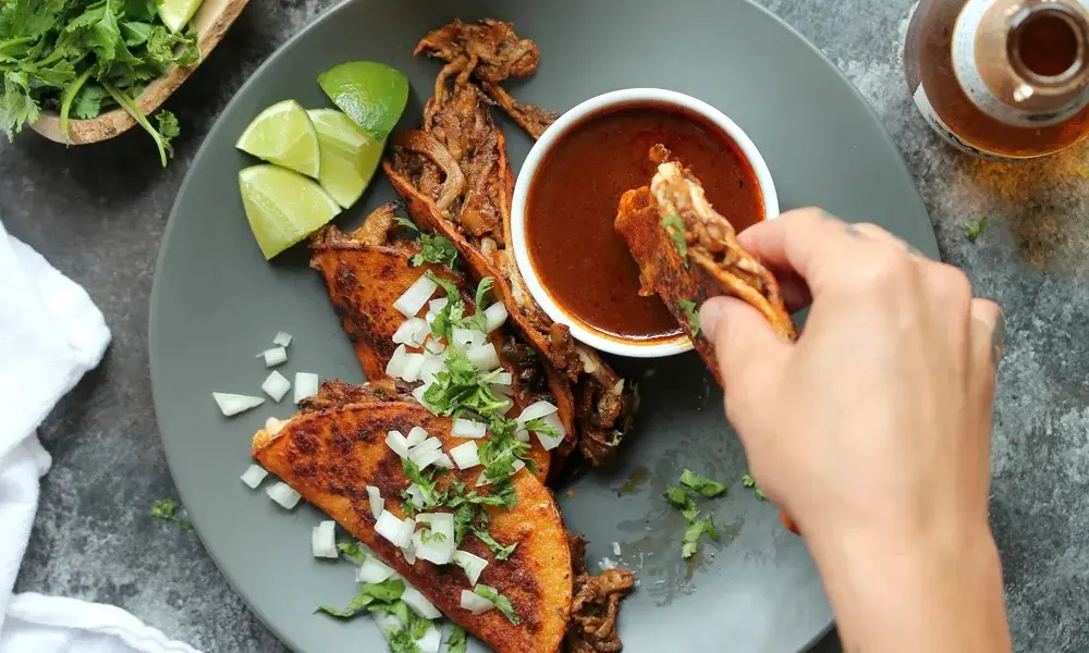 Overhead view of spicy grilled meat tacos with cilantro, diced onion, lime wedges, and red salsa; a hand dips a taco, creating a casual, appetizing mood.