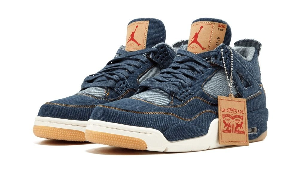 A pair of dark blue denim Air Jordan sneakers with black laces, gum soles, orange Jumpman logos, and a vintage-style hangtag, displayed on white.