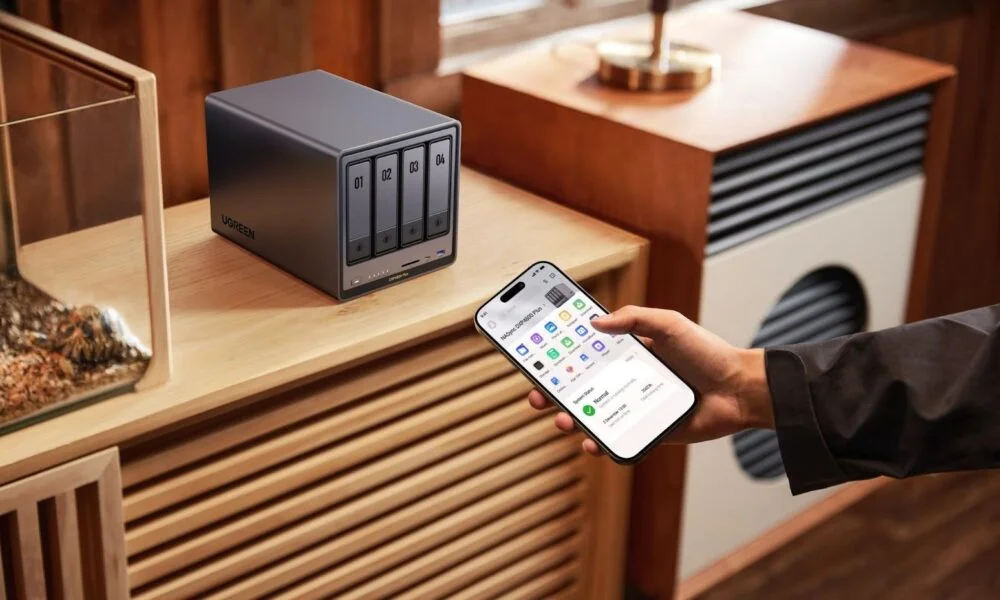 A hand checks a smartphone app beside a compact four-bay network storage device on a wooden cabinet in a warm, modern living room.