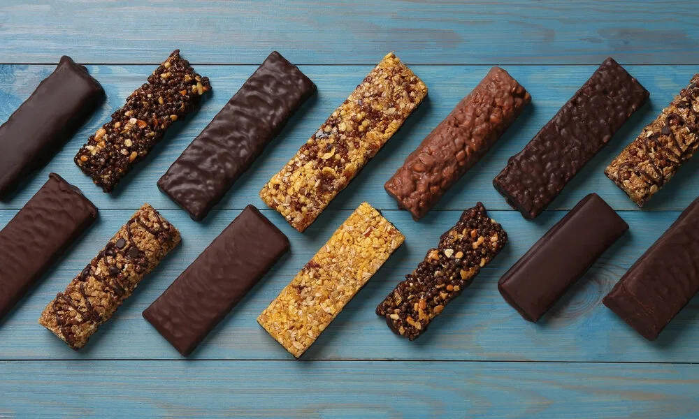 Assorted chocolate and granola energy bars arranged diagonally in rows on a rustic blue wooden surface, showcasing varied textures and toppings.