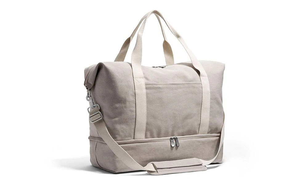 A light beige canvas duffel bag with two handles, a long shoulder strap, front zip pocket, and metal clasps, shown against white; practical and understated.