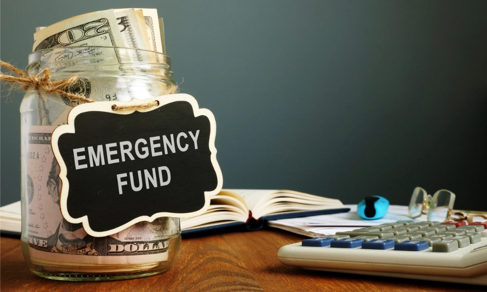 A glass jar filled with cash and labeled “Emergency Fund” sits on a wooden desk beside an open book and calculator, suggesting careful financial planning.