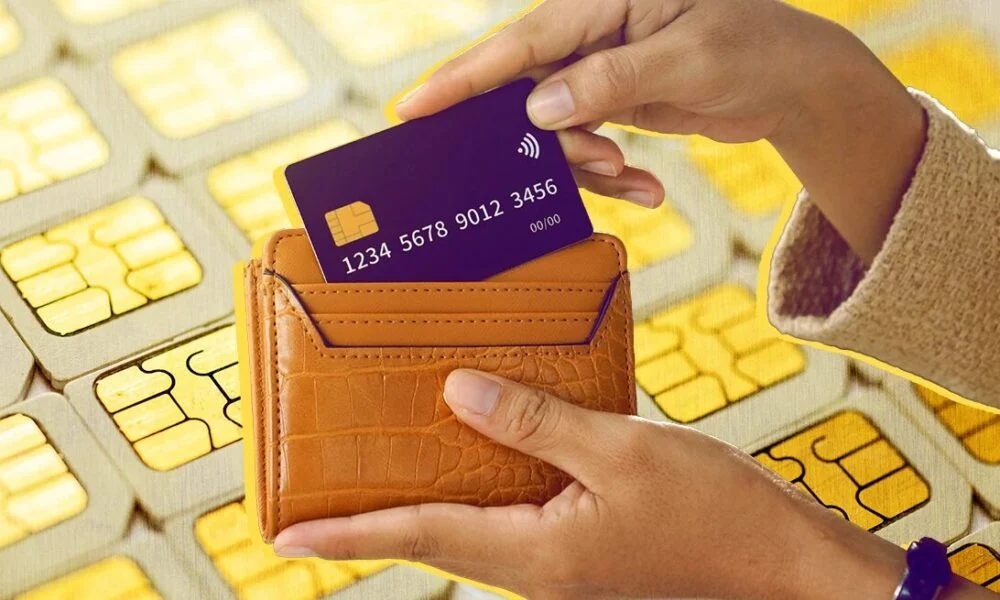 A hand slides a purple bank card into a tan leather wallet