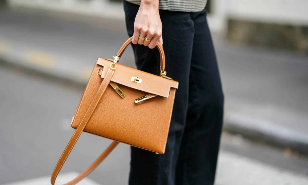 A person in dark trousers carries a structured tan leather handbag with gold hardware and a long shoulder strap, creating a polished street-style look.