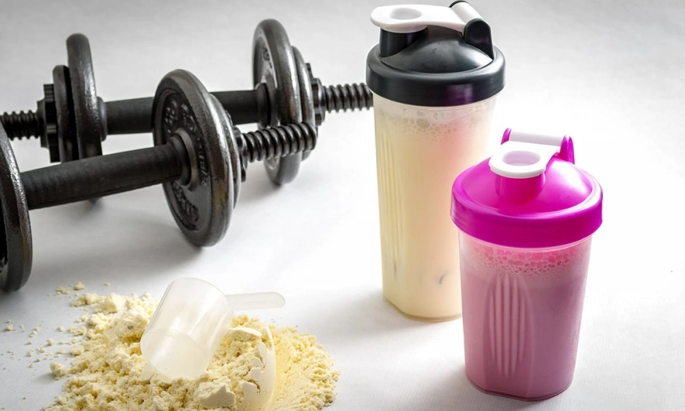 Two protein shaker bottles filled with pale and pink drinks sit beside dumbbells and spilled protein powder, suggesting a post-workout fitness routine.