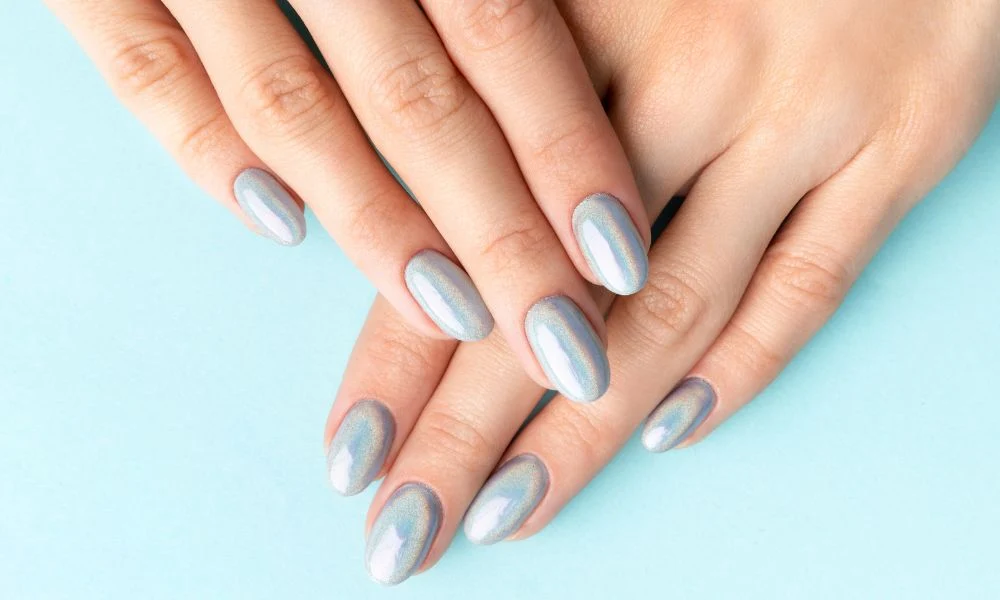 Close-up of manicured hands crossed on a pale blue background, showing long oval nails with shimmering silver-blue polish for an elegant, polished look.