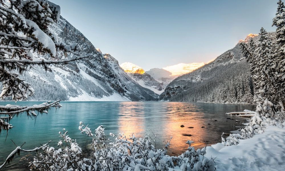 Snow-covered alpine mountains frame a turquoise lake reflecting warm sunset light, with frosted trees and rocks-Prettiest Places in Canada