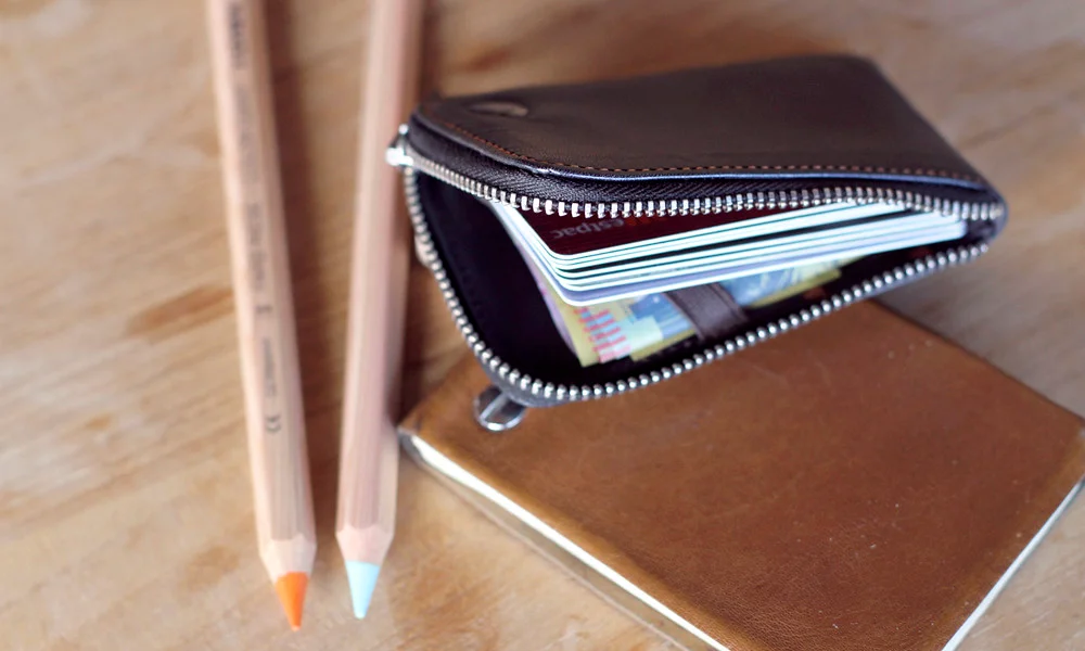 A black zippered wallet holding colorful cards rests on a brown notebook beside two sharpened pencils