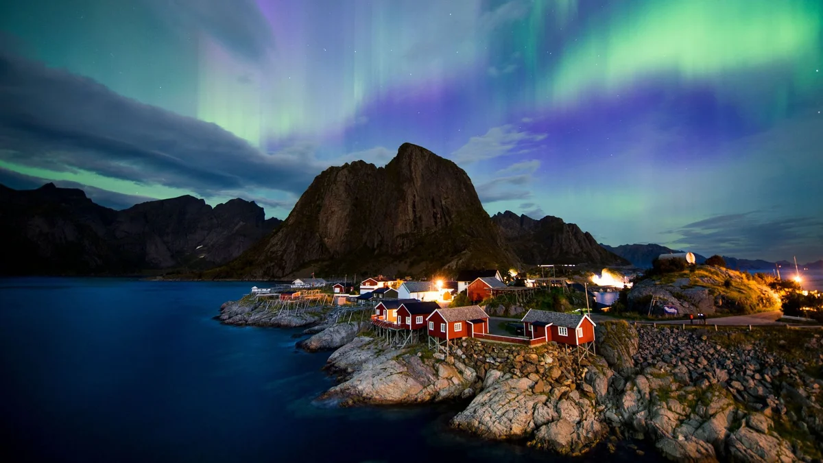 Green and purple northern lights illuminate a remote Norwegian fishing village, its red cabins clustered on rocky shores beneath a towering mountain at night.