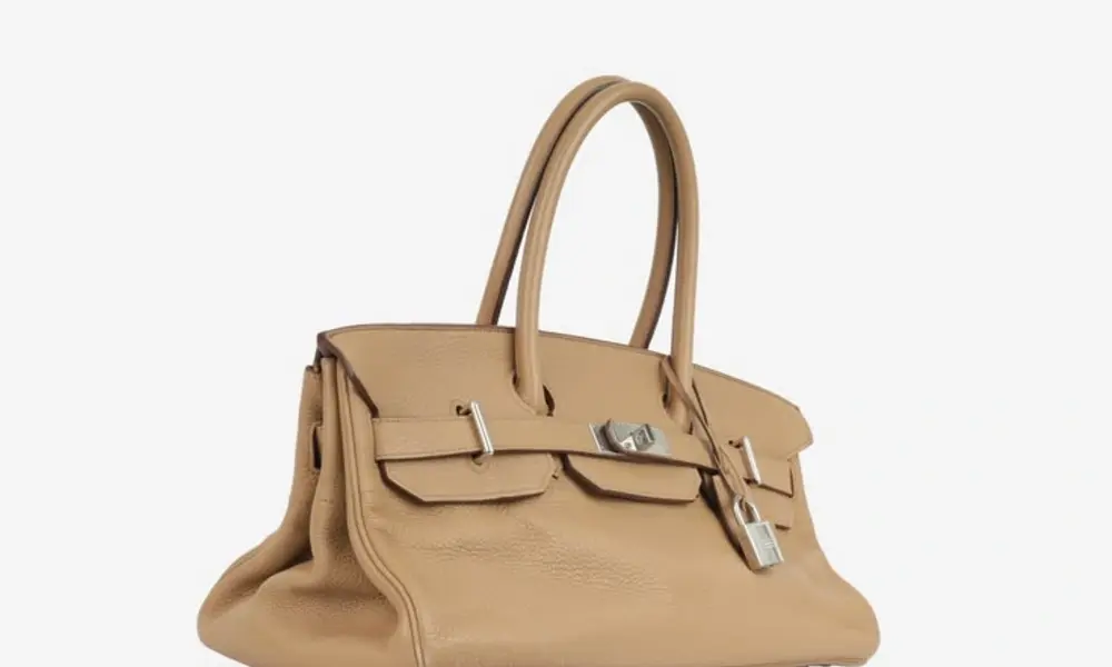 A structured tan leather handbag with rounded top handles, front flap pockets, silver-tone buckles, and a soft slouch at the base, photographed on white.