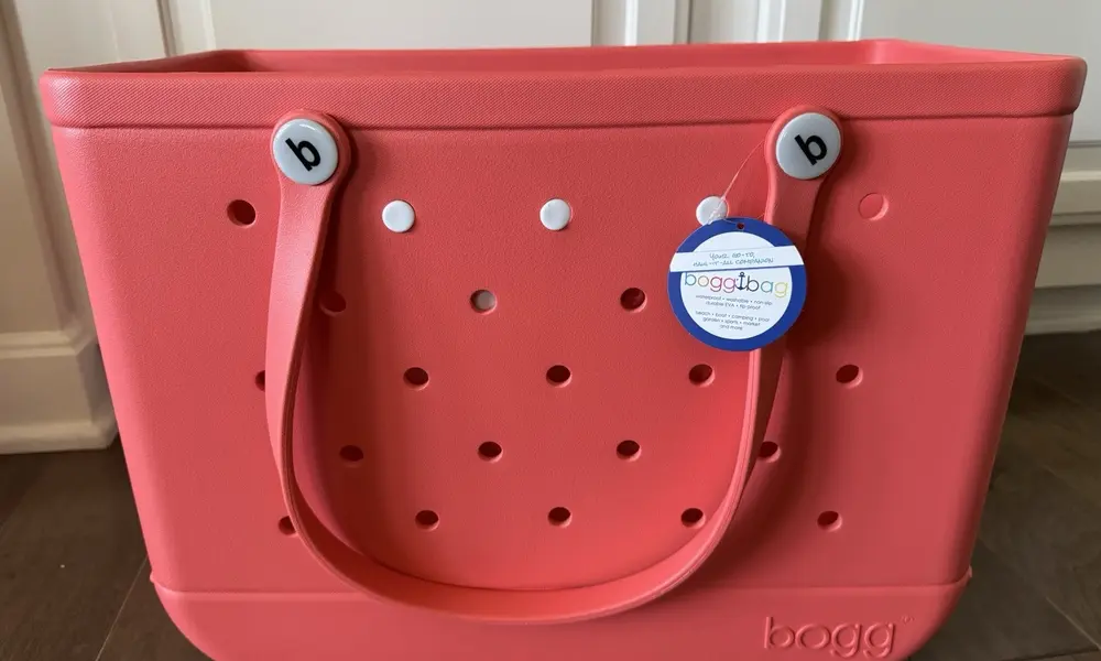 A cheerful coral-pink Bogg tote with perforated sides, matching loop handles, gray button fasteners, and a round logo tag sits indoors.