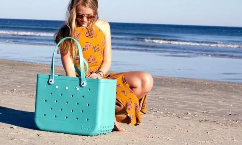 A woman in sunglasses and a yellow floral dress sits on a sunny beach, looking at a large turquoise tote with perforated holes beside the ocean.