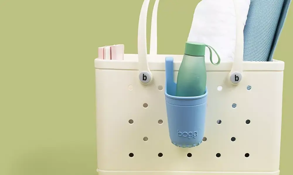 Cream perforated tote bag with a blue hanging cup holding a green water bottle, plus a towel and accessories, against a soft green backdrop.
