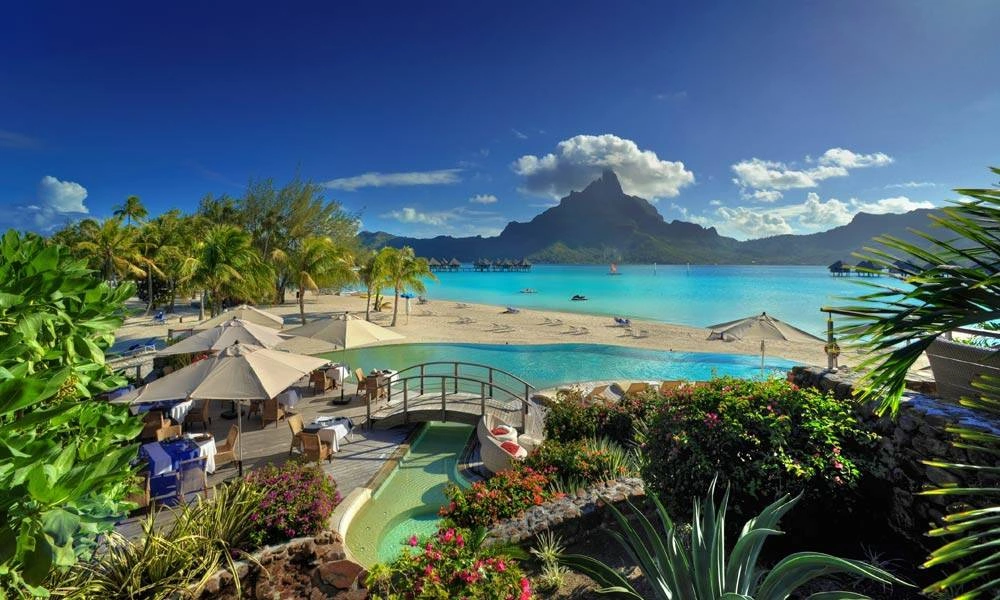 Tropical beach resort with palm trees, thatched umbrellas, turquoise lagoon, gardens, and a dramatic mountain beneath a bright blue sky; serene and inviting.
