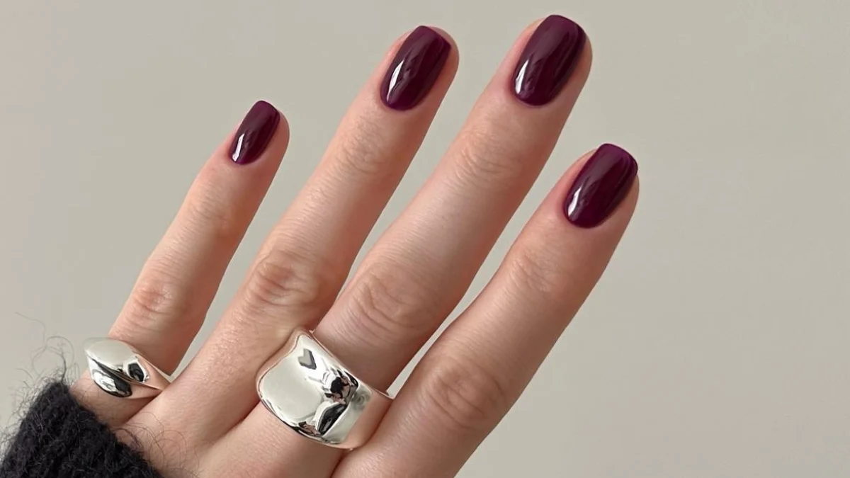 A hand with glossy deep burgundy nail polish wears two silver rings, including a wide sculptural band, against a plain beige background, creating an elegant look.
