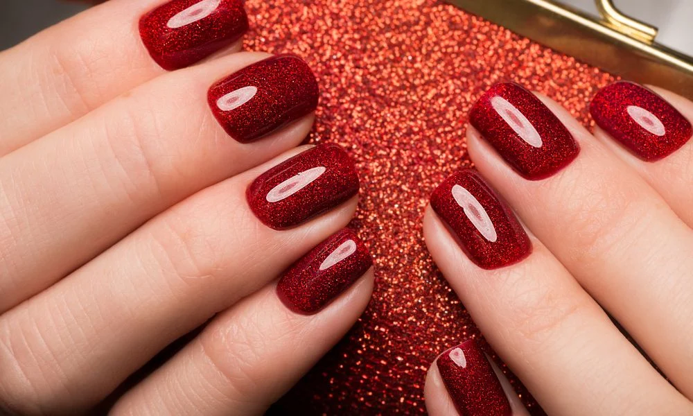 Close-up of hands with glossy deep-burgundy glitter nails resting on a sparkling red surface, creating a glamorous, festive beauty look.