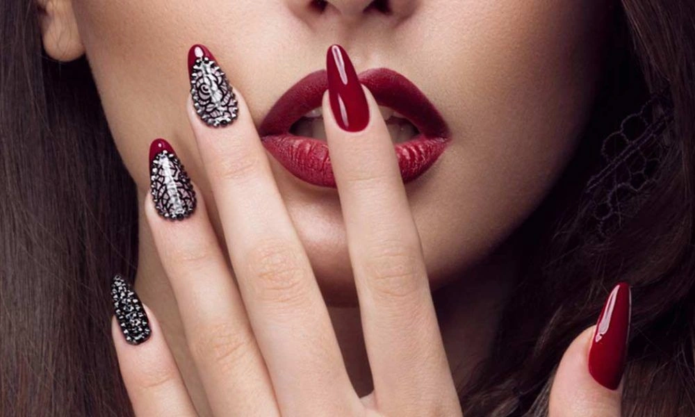 Close-up beauty portrait of a woman with glossy burgundy lips and matching long nails, including black-and-white patterned accents, posed over her mouth for a bold, glamorous mood.