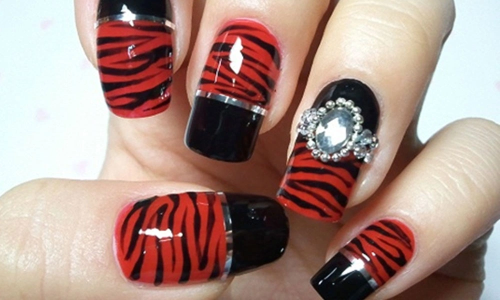 Close-up of manicured nails with glossy red-and-black tiger stripes, black tips, and a sparkling rhinestone accent, creating a bold, glamorous look.