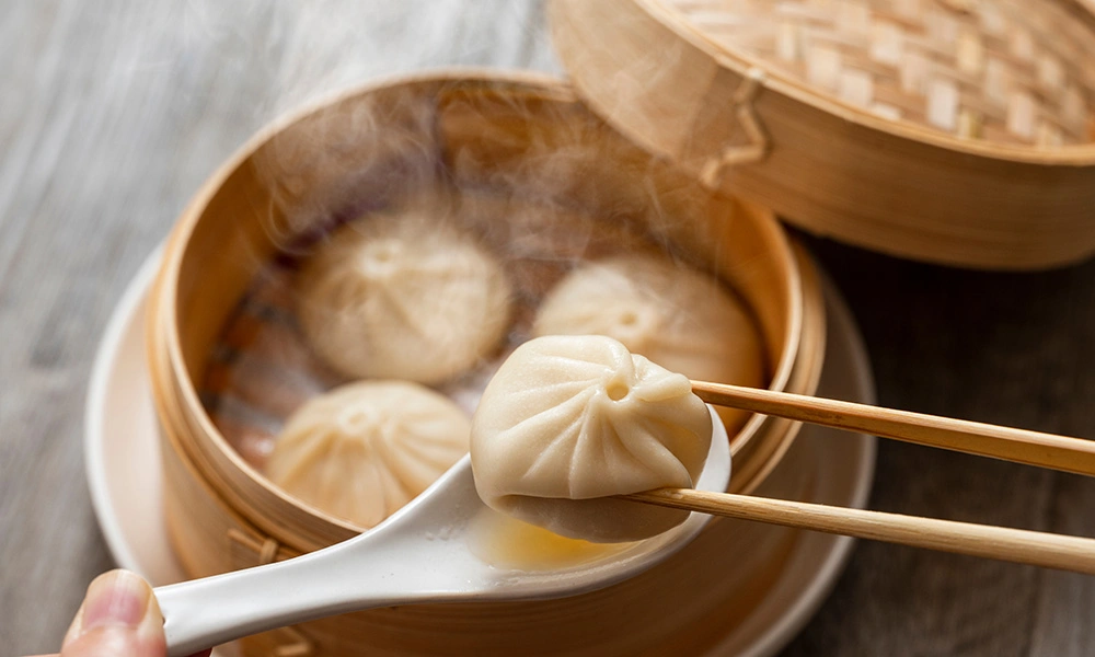 A steaming soup dumpling rests on a white spoon held by chopsticks above a bamboo steamer of dumplings, evoking warm, inviting comfort food.
