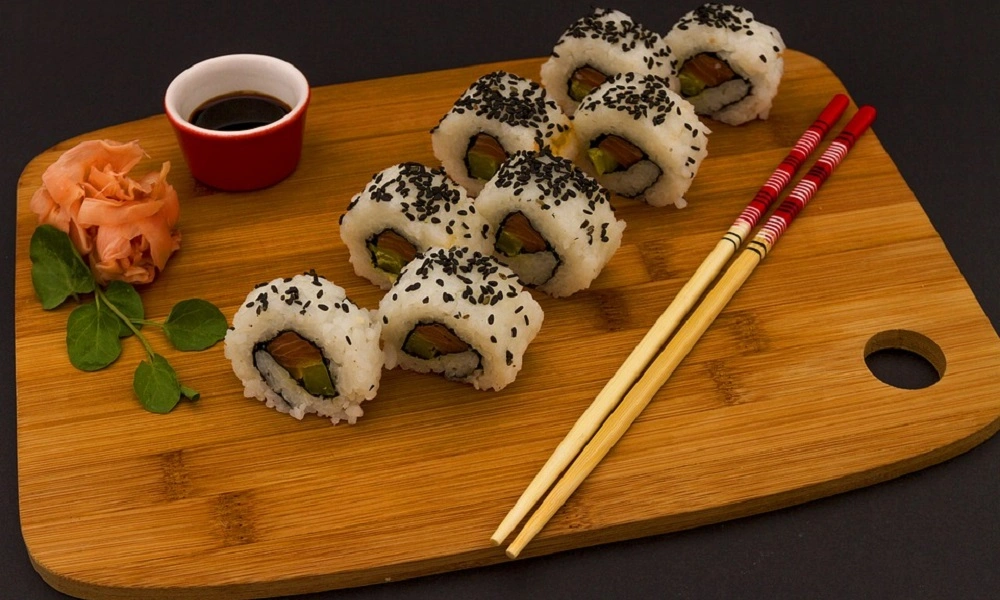 A wooden board holds sesame-topped sushi rolls, pickled ginger, green leaves, soy sauce, and red-tipped chopsticks, arranged neatly for a calm meal.