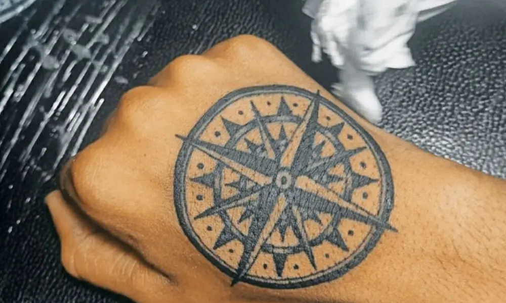 Close-up of a hand being tattooed with a bold circular compass design, featuring a central star and geometric patterns; the scene feels focused and calm.