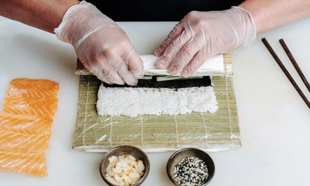 Gloved hands roll sushi rice and seaweed in a bamboo mat, with sliced salmon, bowls of toppings, and chopsticks nearby; a calm, careful food-preparation scene.