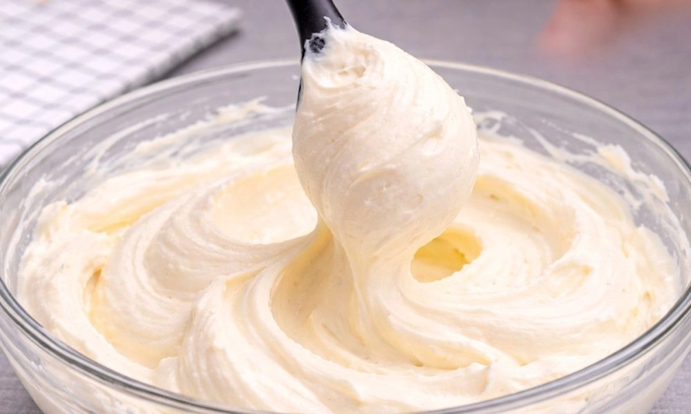 A spoon lifts a soft peak of creamy pale-yellow frosting from a glass bowl, its smooth swirls suggesting a rich, freshly whipped dessert topping.