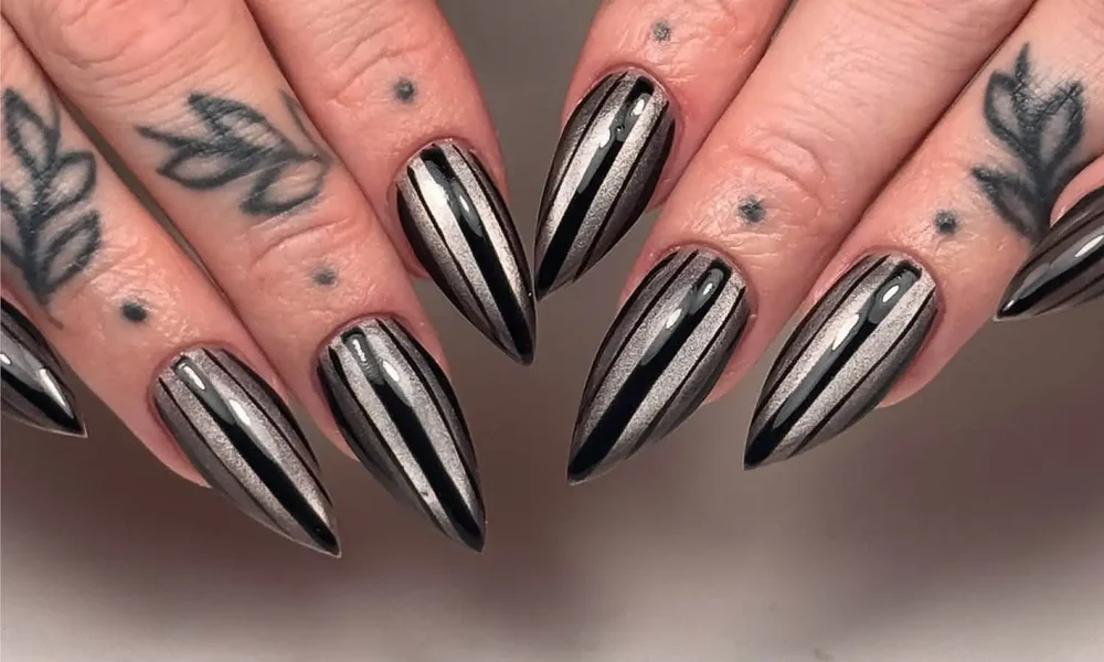 Close-up of tattooed fingers with long pointed black-and-silver chrome nails, arranged against a neutral background for an edgy, dramatic look.
