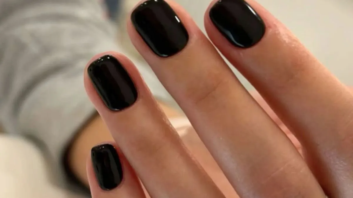 Close-up of a hand with glossy black nail polish, fingers gently curved against a soft gray background, creating a sleek, polished look.