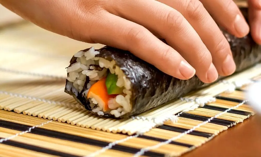 Hands gently roll a nori-wrapped sushi roll on a bamboo mat, with rice, carrot, and green vegetable filling visible in this calm cooking scene.