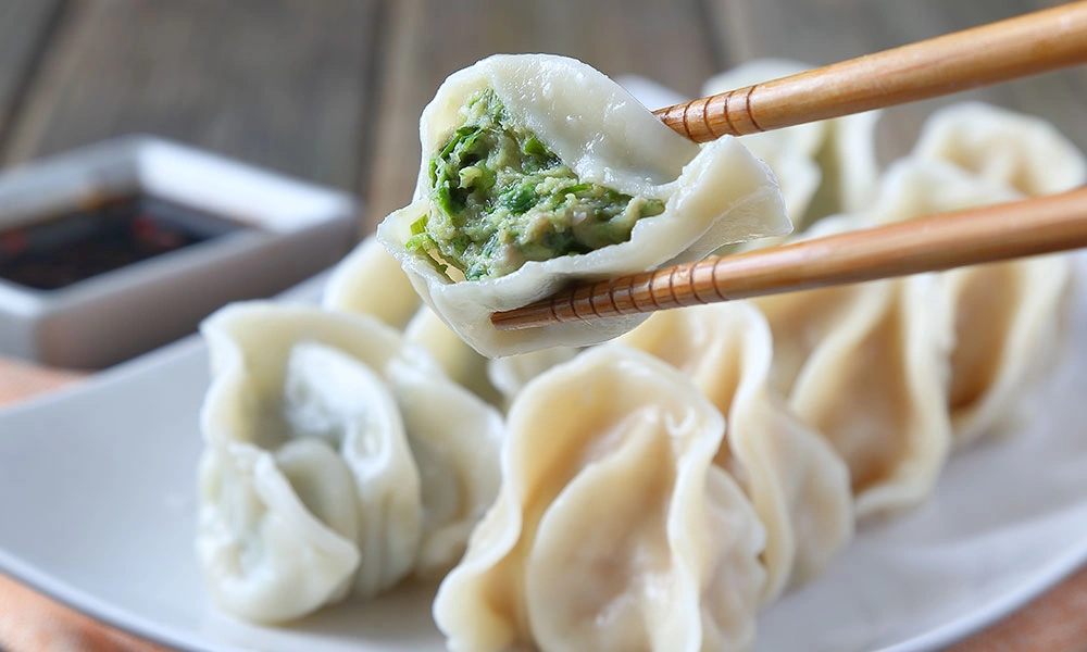 Chopsticks hold a steamed dumpling split open to reveal green vegetable filling, above a plate of dumplings with dipping sauce in the background.