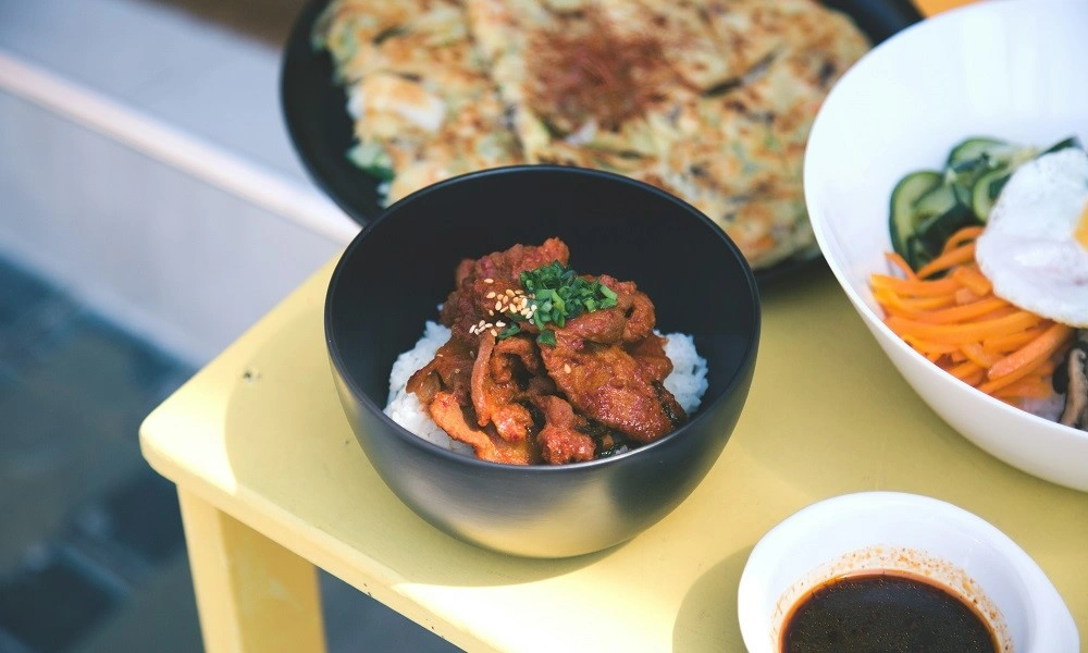 A black bowl of spicy red meat over white rice, topped with sesame and herbs, sits on a yellow table beside a pancake-Best Chili Recipe