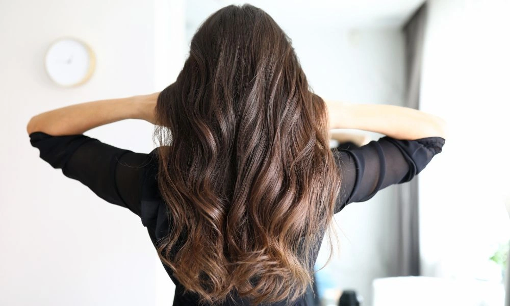 A woman seen from behind gently runs her hands through long, glossy brown waves in a bright, minimalist room, conveying a relaxed hair-care moment.