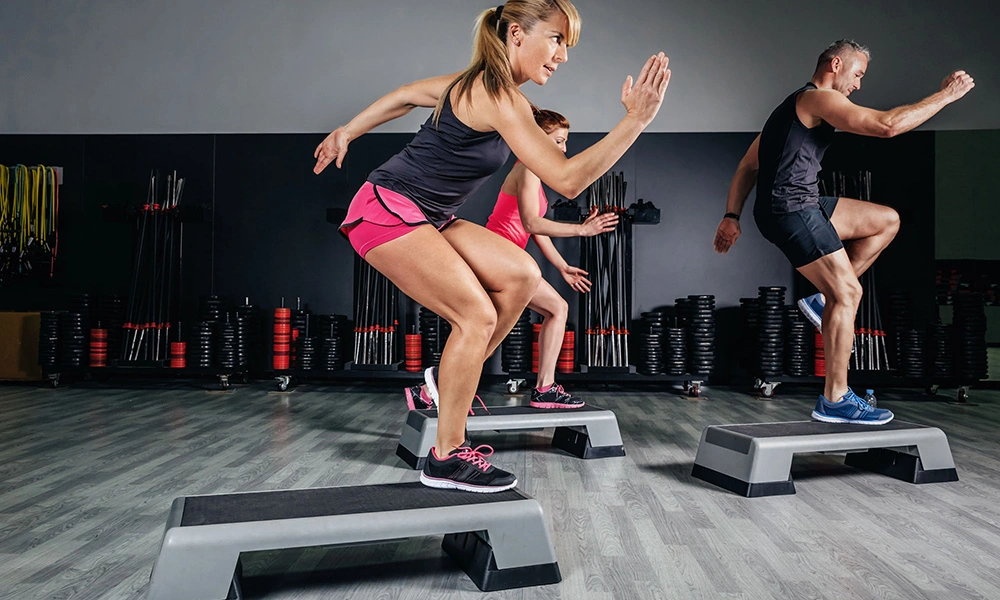 Three adults perform energetic step-ups on aerobic platforms in a gym, focused on their workout as fitness equipment lines the dark wall.