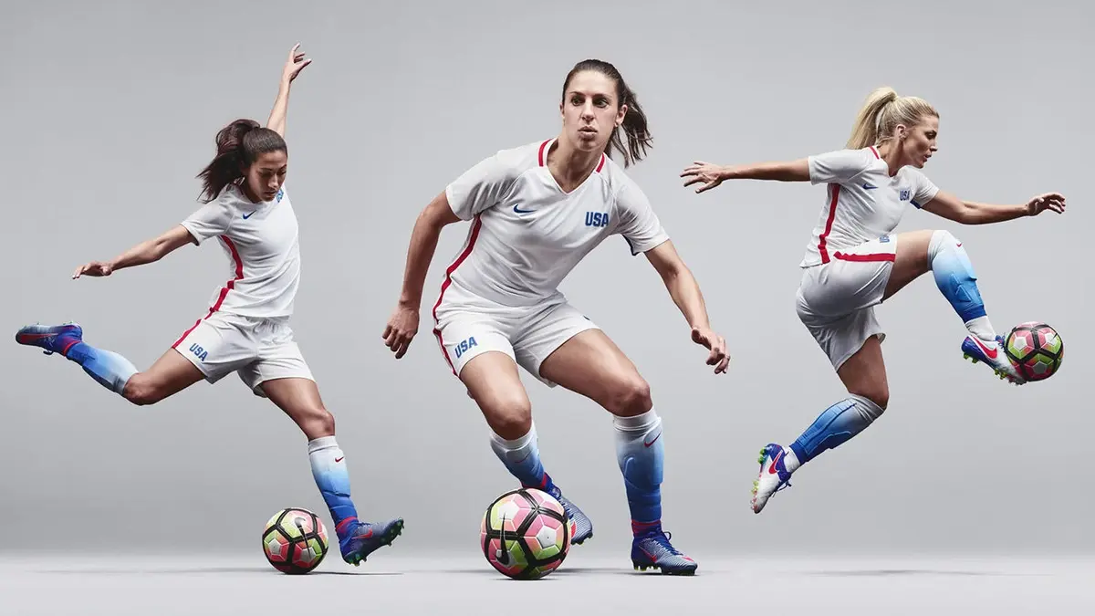 Three female soccer players in white USA uniforms control colorful balls mid-drill against a gray studio backdrop, captured in energetic poses that convey athletic focus.