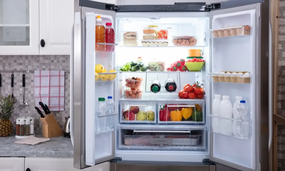 An open, brightly lit refrigerator neatly stocked with fresh fruit, vegetables, drinks, and dairy in a modern kitchen, conveying freshness and abundance.